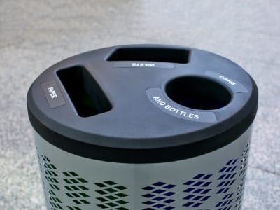 top angled view of a triple stream waste and recycling container with a metal body and black plastic lid on a city sidewalk