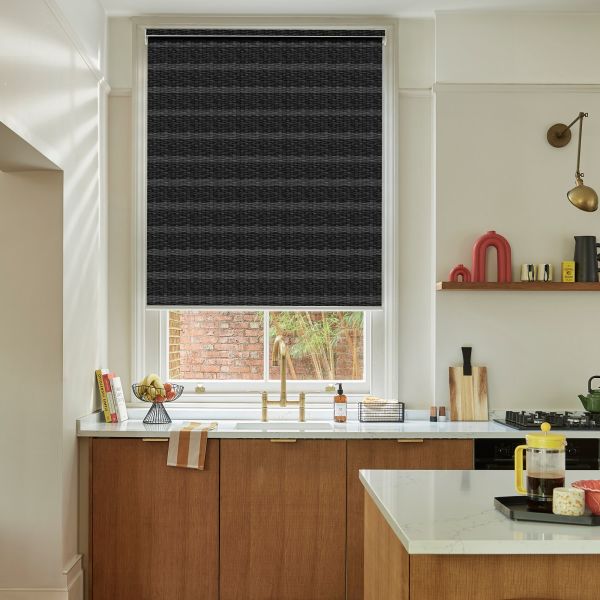 Dark charcoal woven roller blind fully lowered across window, showing horizontal textured weave and silver top rail; blocking light over a kitchen sink with brass tap, wood cabinets and marble countertop.