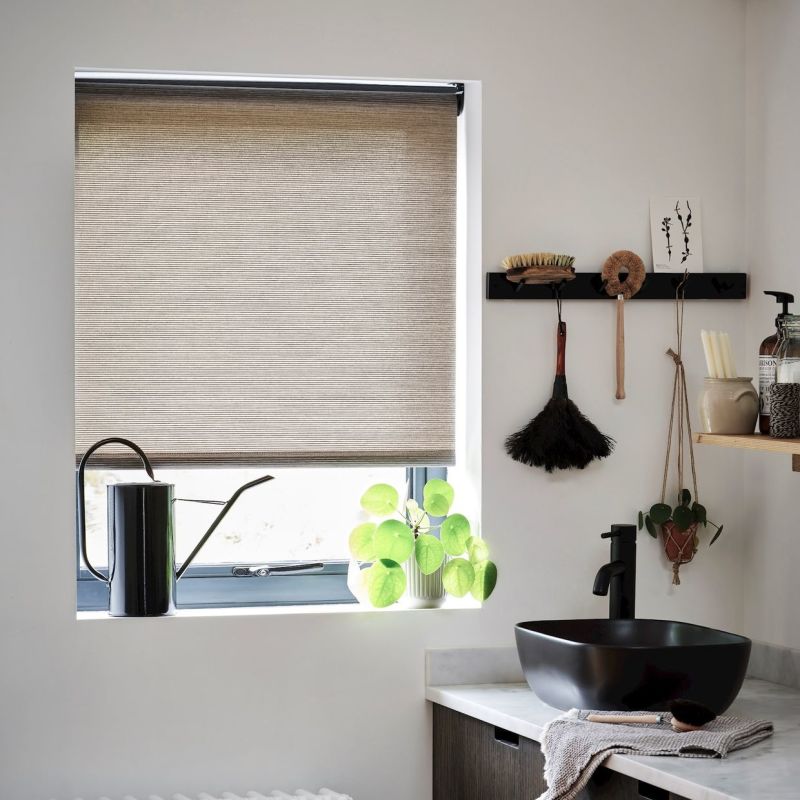 Beige fabric roller blind lowered over a recessed window, filtering daylight; foreground shows a black watering can, potted roundâ€‘leaf plant, black vessel sink and hanging brushes on a white wall.