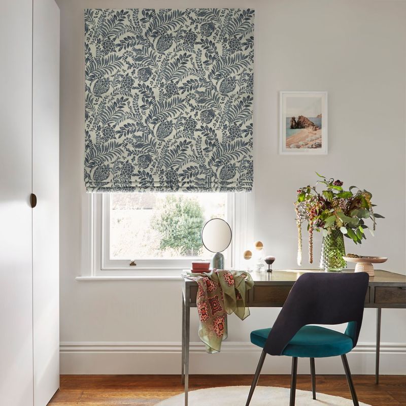 Roman blind with blue botanical pattern covering window — lowered, partially filtering daylight; small pale-walled home office with wooden desk, teal chair, vase of flowers, mirror and framed seascape.