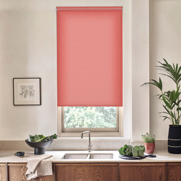 Coral roller blind covers a kitchen window, partially lowered over a sink; stainless faucet centered, countertop holds bowls of leafy greens, potted plants, and a framed sketch on the wall.