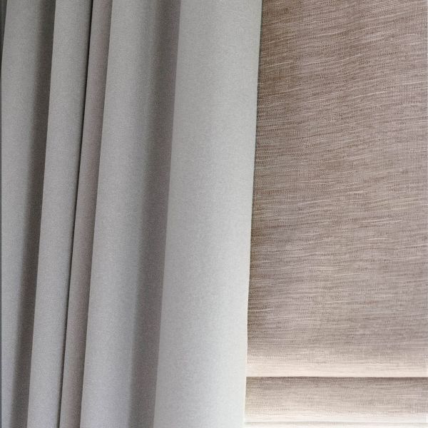 Grey pleated blackout curtain at left and beige textured Roman blind at right; both lowered, the curtain drawn closed and the blind filtering diffuse daylight in a close-up interior view.