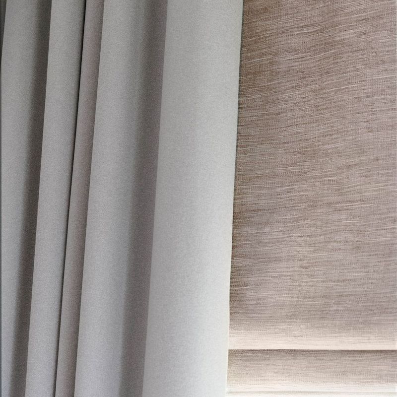 Grey pleated blackout curtain at left and beige textured Roman blind at right; both lowered, the curtain drawn closed and the blind filtering diffuse daylight in a close-up interior view.