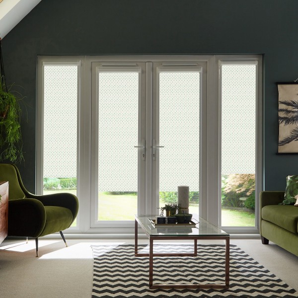 Double glass patio doors stand closed with white patterned roller blinds diffusing daylight, framing a living room with a glass coffee table, chevron rug, green armchairs, and potted plants.