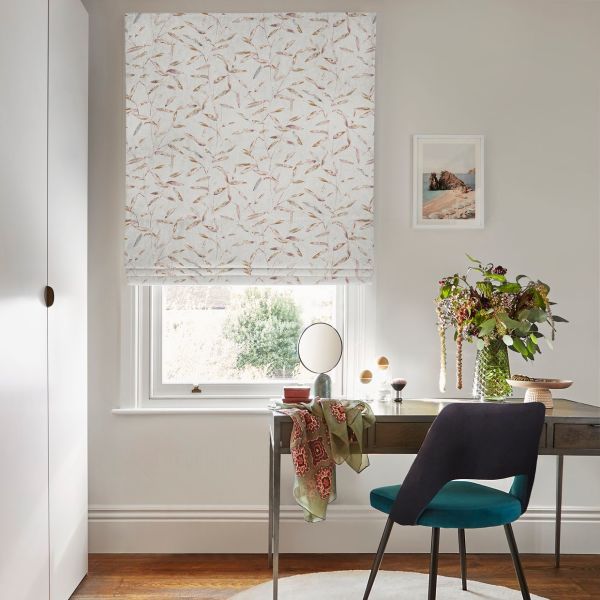 A patterned Roman blind (white with pale leaves), fully lowered and filtering daylight; over a window above a desk with mirror, vase of flowers and teal chair in a study.