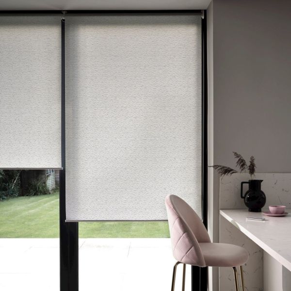 White semi-opaque roller blinds covering floor-to-ceiling black-framed glass doors, lowered to block view; soft daylight filters around edges into a modern kitchen nook with pink barstool and countertop.
