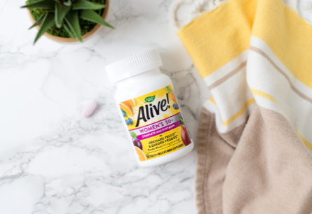 A bottle of Alive Women's 50 Plus Multivitamins laying on a marble countertop in between a kitchen towel and a potted succulent.