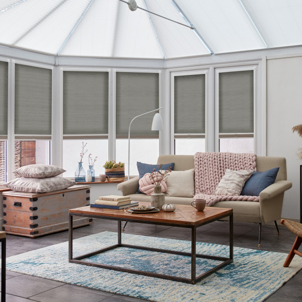 Pleated cellular blinds across tall bay windows, lowered to mid-height and filtering daylight; modern conservatory living room with vaulted glass roof, beige sofa, wooden coffee table and blue rug.