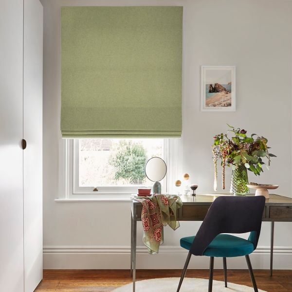 Green fabric Roman blind, lowered with soft folds at bottom, filtering daylight through the uncovered lower pane; above a wooden desk in a bright office with chair and vase.