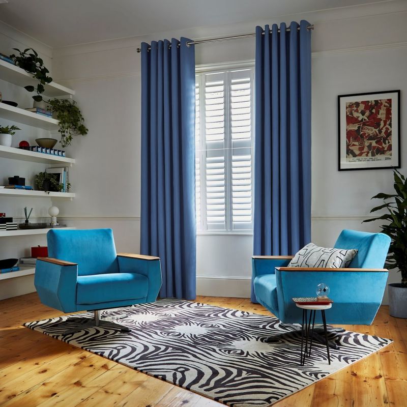 Blue grommet-top curtains flank a window with white plantation shutters, hanging open and filtering daylight; mid-century turquoise armchairs face each other on a zebra rug in a bright living room.