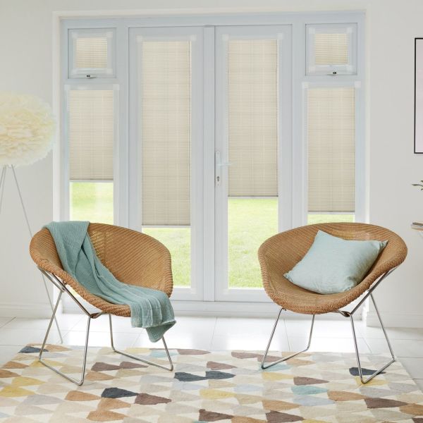Beige horizontal-slat blinds fitted inside French doors and sidelights, lowered and closed, filtering soft daylight; modern living area with two rattan chairs, pastel cushions, patterned rug and lawn view.