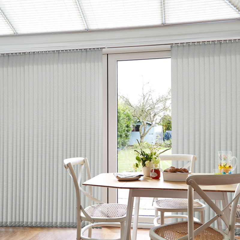Light-gray vertical blinds, mostly closed with a narrow central gap filtering daylight; wooden dining table and wicker chairs in a bright conservatory-style kitchen, garden and shed visible outside.