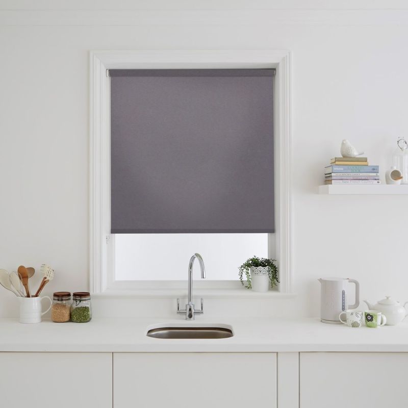 A grey roller blind fully lowered over a rectangular window, blocking light; set above a white kitchen sink with chrome faucet, potted plant, jars, kettle and minimalist shelving.