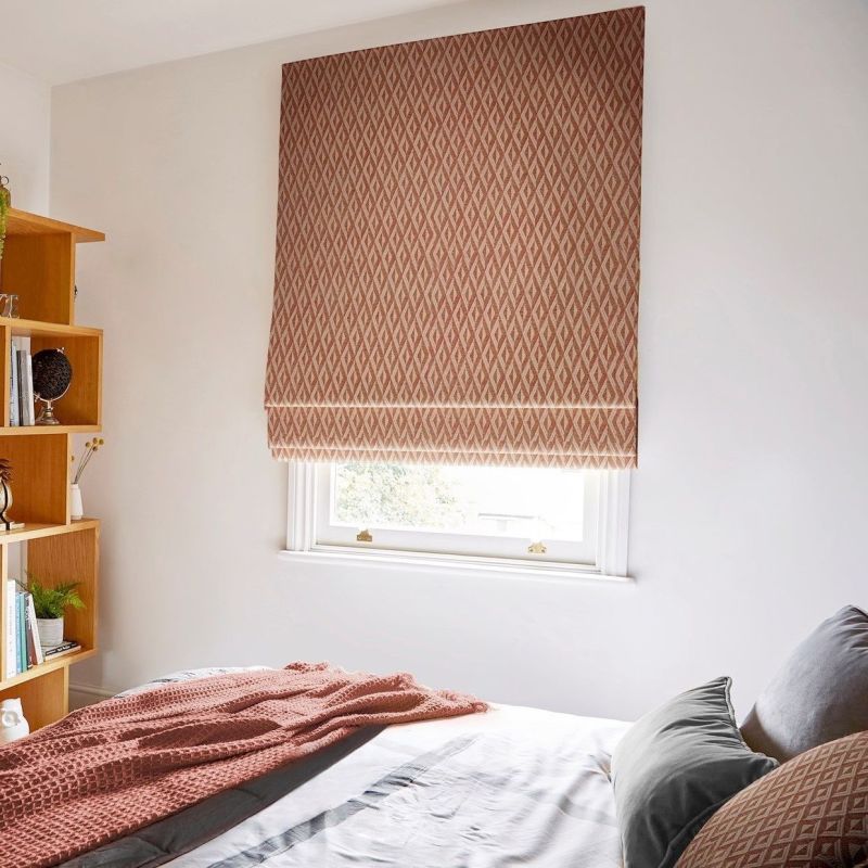 Geometric-patterned fabric Roman blind lowered over a small sash window, softly filtering daylight; set in a bright bedroom with a bed, textured coral throw and wooden shelving.