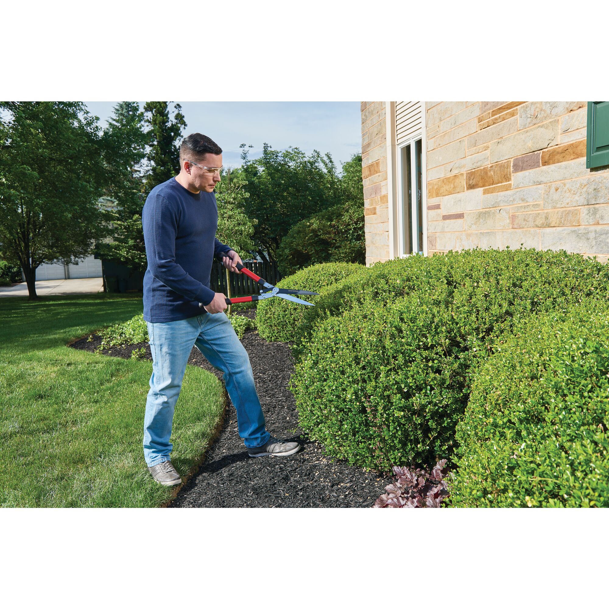 Hedge shears with compound action blade being used by a person to trim a hedge outdoors.