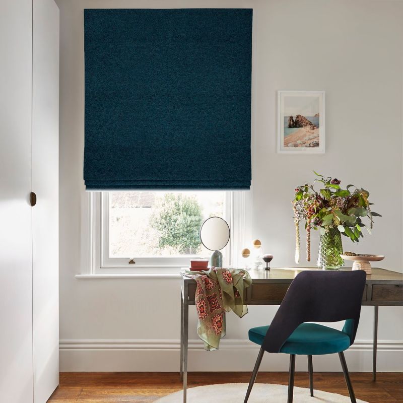 Dark teal Roman blind, lowered with soft horizontal folds, filtering daylight over a white window in a bright home office; wooden desk with vase of flowers and a teal chair.