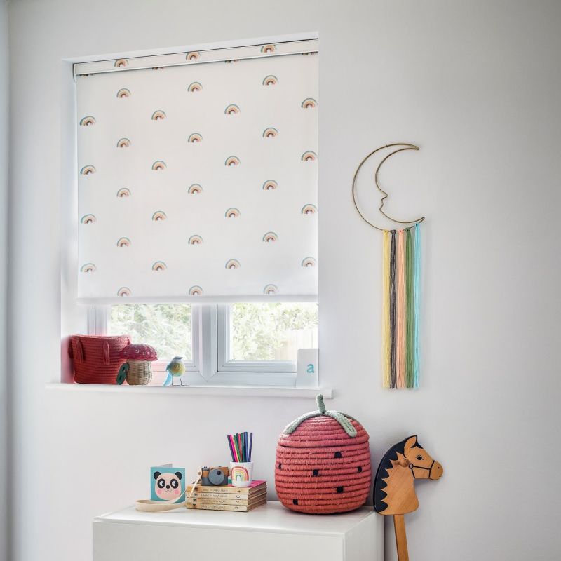 White roller blind printed with small rainbows lowered over the window, filtering soft daylight; nursery shelf and dresser display toys, a strawberry basket and crescent wall-hanging with pastel tassels.

a