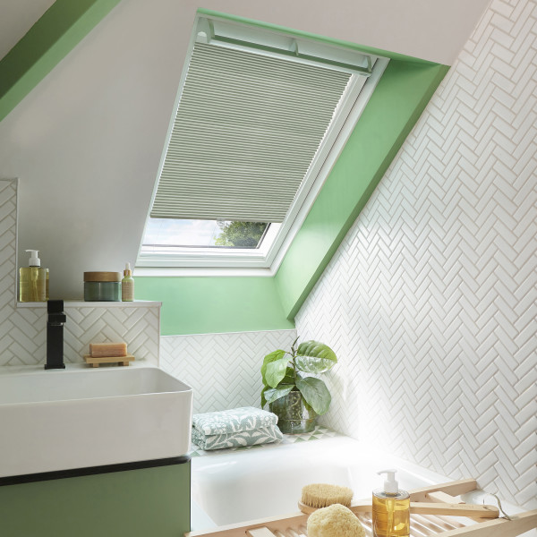 Pleated skylight blind (white), partially lowered over a roof window, filtering daylight. In an attic bathroom with mint-green window frame, white herringbone tiles, sink, bathtub, plant and toiletries.