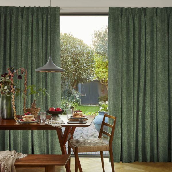 Floor-to-ceiling green textured pleated curtains hang in soft folds, slightly parted to reveal a sunlit garden; a dining table set for breakfast with pancakes, pendant lamp and wooden chairs.