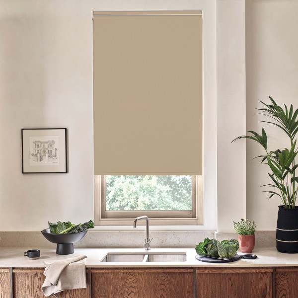 Beige roller blind covers most of a window, partially lowered above a kitchen sink and faucet, set on a countertop with bowl of greens, framed sketch, and potted plants.