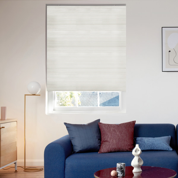 Off-white pleated cellular blind, lowered across the window with a small bottom gap, filtering daylight; positioned above a navy sofa with throw pillows, floor lamp, sideboard, and minimal living-room decor.