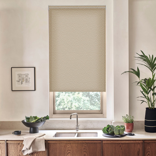 Beige patterned roller blind lowers over a kitchen window, filtering light; stainless faucet and sink sit below on a countertop with bowls of leafy greens, potted plants, and a framed sketch.
