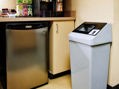 single trash bin with a black sloped lid and restrictive opening in an office break room beside a mini fridge and coffee station