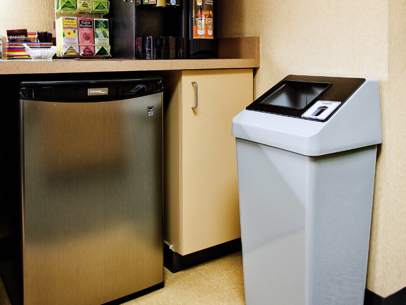 single trash bin with a black sloped lid and restrictive opening in an office break room beside a mini fridge and coffee station