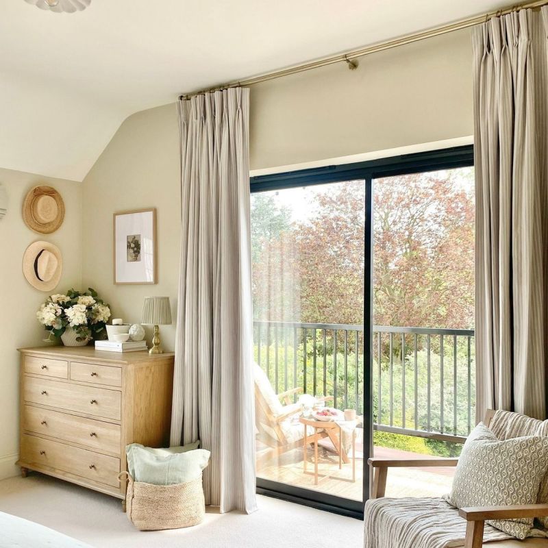 Floor-to-ceiling pleated beige curtains on a brass rod, parted to reveal a sliding glass door; filtering soft daylight into a neutral bedroom corner with wooden dresser, balcony chairs and leafy view.