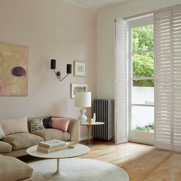 White plantation shutters on a French door, panels partly open, filtering daylight and framing a leafy garden; softly lit living room with beige sofa, round coffee table, and wooden floor.