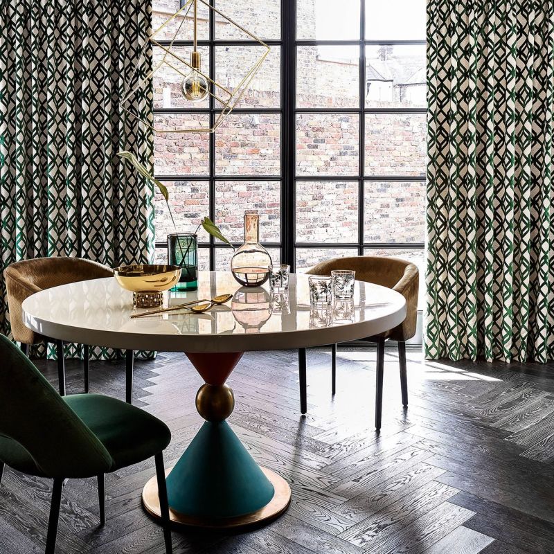 Green-and-black geometric full-length drapes, drawn to sides, framing a black-grid window and filtering daylight; dining area with round table, glassware, pendant light, and dark herringbone floor.