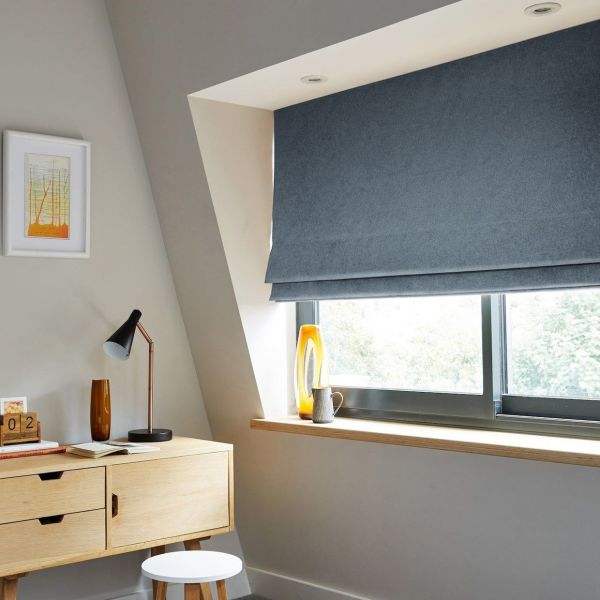 Blue Roman Roman blind lowered in layered folds, covering the upper casement window and partially filtering daylight; small sloped‑ceiling home office with light wood desk, lamp, stool, vase on sill. 02 JUNE