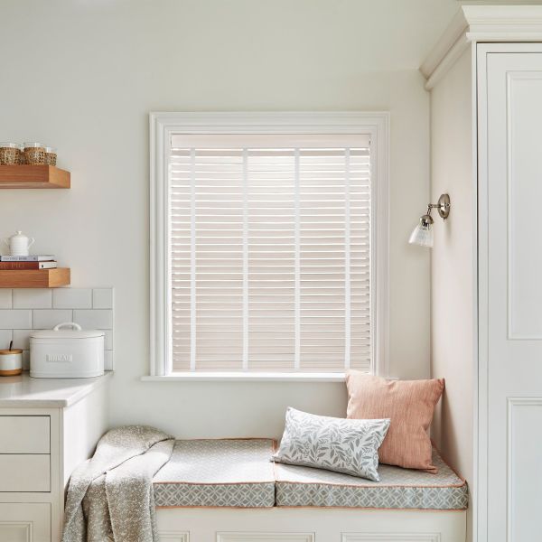 White horizontal slatted blinds within a painted window frame, closed and filtering soft daylight; kitchen nook with cushioned bench, throw and pillows, floating shelves, bread box and neutral decor.
BREAD