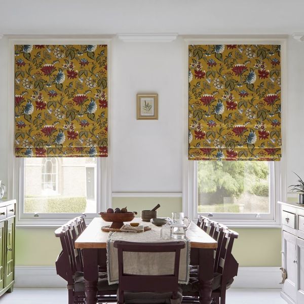 Two mustard-yellow floral Roman blinds, lowered over adjacent windows, patterned with red and blue flowers, filtering daylight into a dining room with wooden table, chairs and garden view.