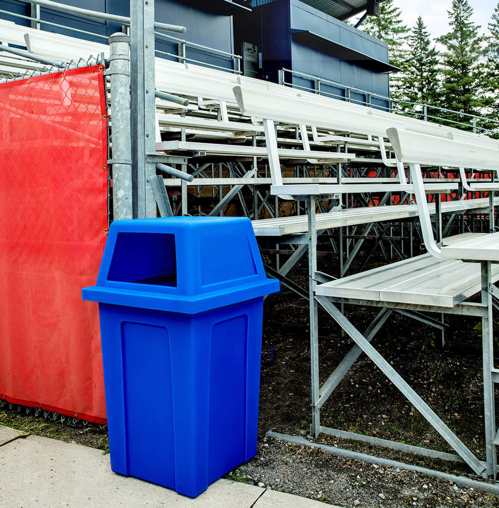 large capacity blue recycling bin with a rain hood lid sitting beside bleachers at a baseball field