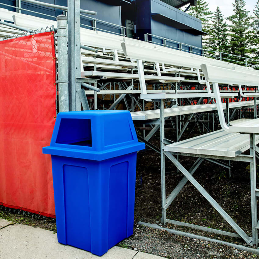 large capacity blue recycling bin with a rain hood lid sitting beside bleachers at a baseball field