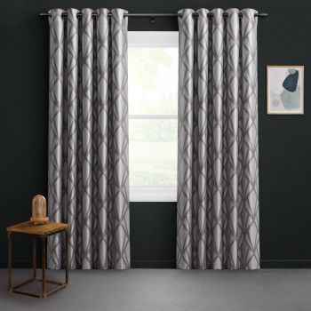 Grommet-top silver-gray curtains with dark geometric lattice, parted at center over a window, filtering soft daylight; dark charcoal walls, small wooden side table with glass-dome lamp, framed abstract art.