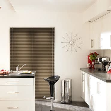 Horizontal Venetian blinds covering a recessed window, fully lowered and closed, filtering light; modern white kitchen with glossy countertops, chrome stool, trash bin, wall clock and vase of red tulips.