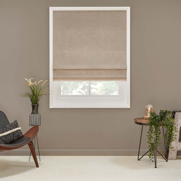 Beige fabric Roman shade, partially lowered with soft horizontal folds, filtering daylight through a lower gap; set on a taupe wall in a neutral living room with mid-century chair, plants, and side table.
