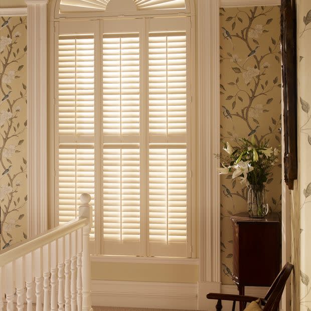 White plantation shutters covering a tall arched window, closed and filtering warm daylight; positioned on a stair landing with floral wallpaper, a wooden banister, side table and vase of lilies.