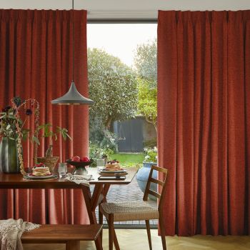 Heavy rust-red pleated curtains, pulled to each side and parted to reveal a glass door, filtering soft daylight over a dining table set for breakfast and a green backyard.