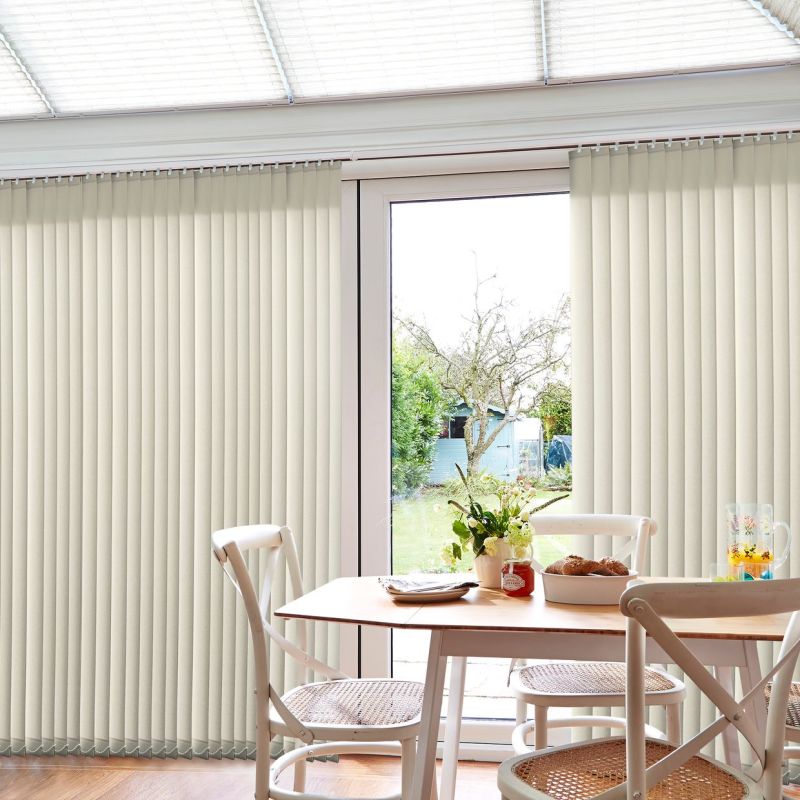 Vertical fabric blinds, cream-colored and mounted at the top, panels closed at the sides and parted centrally revealing a glass patio door; dining table with chairs, breakfast items, and garden beyond.