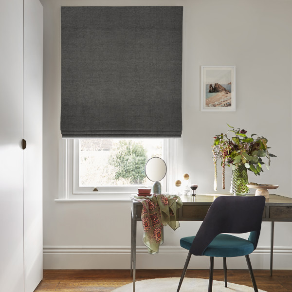 Dark grey Roman blind lowered over window, mostly closed and filtering daylight with an open strip at bottom; above desk with mirror, teal chair, floral vase in bright room.
