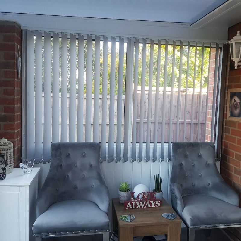Vertical white blinds cover a wide conservatory window, slats slightly angled and filtering daylight. Two grey tufted chairs flank a wooden side table amid brick walls and a fenced garden view.

LOVE ALWAYS