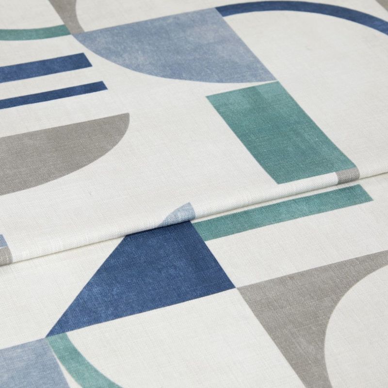 Roman blind with white background and blue, teal, and gray geometric shapes, folded in horizontal pleats; fabric lies flat, showing printed semicircles, rectangles and triangles in soft indoor lighting.