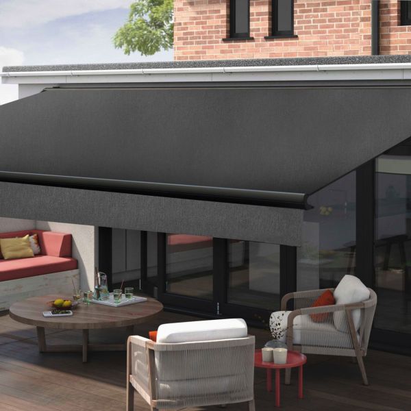 Dark charcoal retractable awning, mounted above glass doors with roller bar, extended and shading the seating area; outdoor wooden-deck patio attached to a brick house, cushioned chairs and round table.