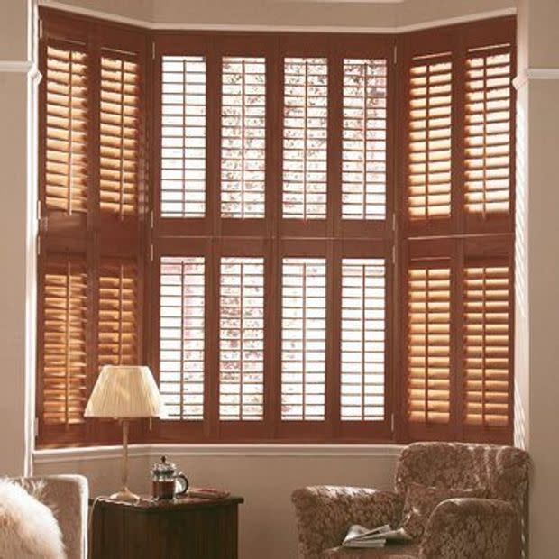 Bay window covered by wooden plantation shuttersâ€”multi-panel, upper and lower sections with tilted slats; filtering warm daylight into a living room with upholstered armchairs, lamp, and side table.