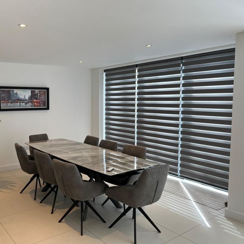 Layered zebra roller blinds cover full-height sliding windows, lowered with alternating opaque and sheer bands filtering daylight; dining room features marble table, eight gray upholstered chairs and recessed ceiling lights.