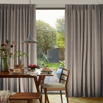 Floor-length gray pleated curtains with pinch pleats, pulled aside to frame a glass door and admit daylight; wooden dining table set for breakfast, pendant light, garden and shed beyond.