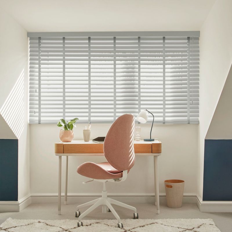 White horizontal venetian blinds cover a wide dormer window, slats tilted to filter sunlight and cast stripes; a small attic study with a wooden desk, pink swivel chair and lamp.